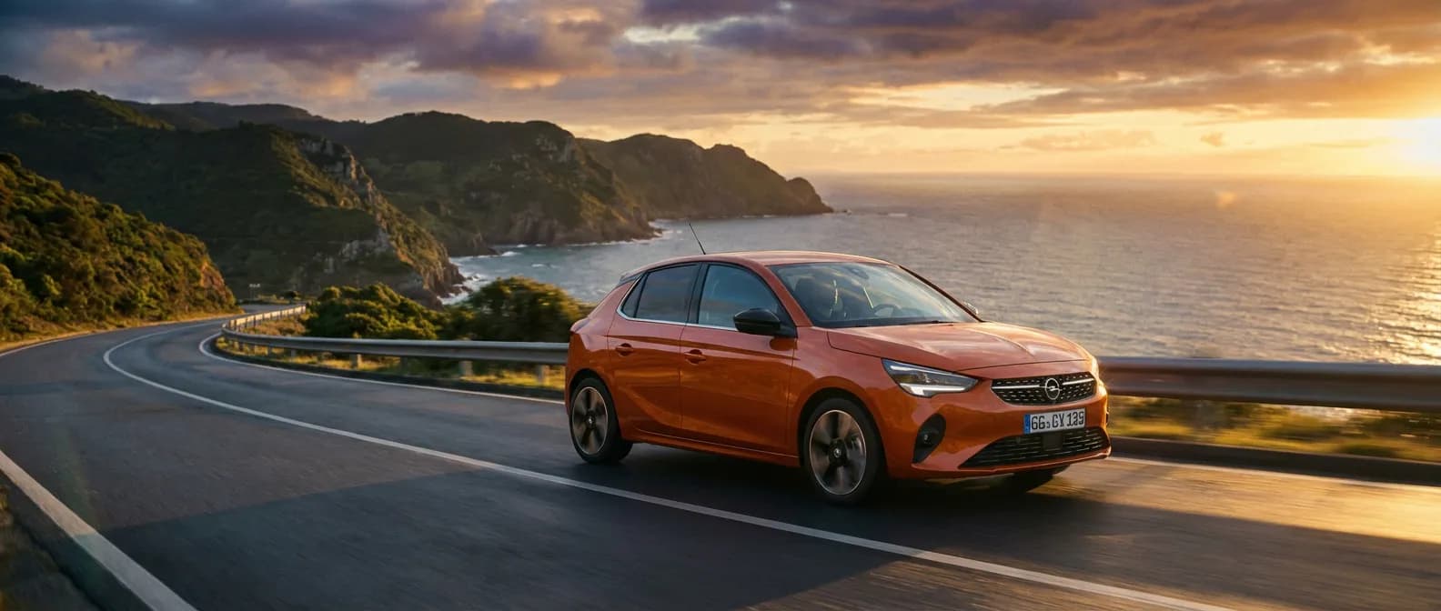 Opel Corsa neuve rouge roulant sur une route côtière au coucher du soleil, paysage épique.
