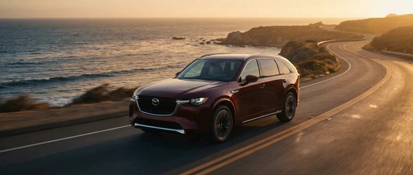Mazda CX-90 SUV hybride premium roulant sur une route côtière au coucher du soleil